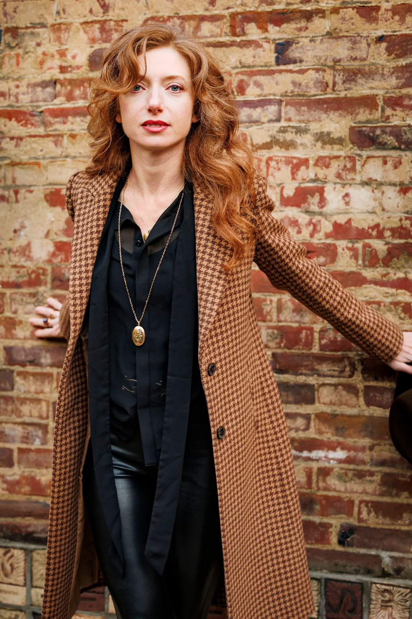 Olya in a houndstooth coat, standing against a weathered brick wall